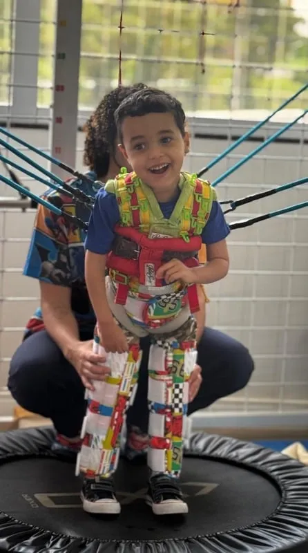Criança sorrindo em pé sobre uma cama elástica durante sessão de fisioterapia com veste terapêutica, apoiada por uma terapeuta na Baby Kids